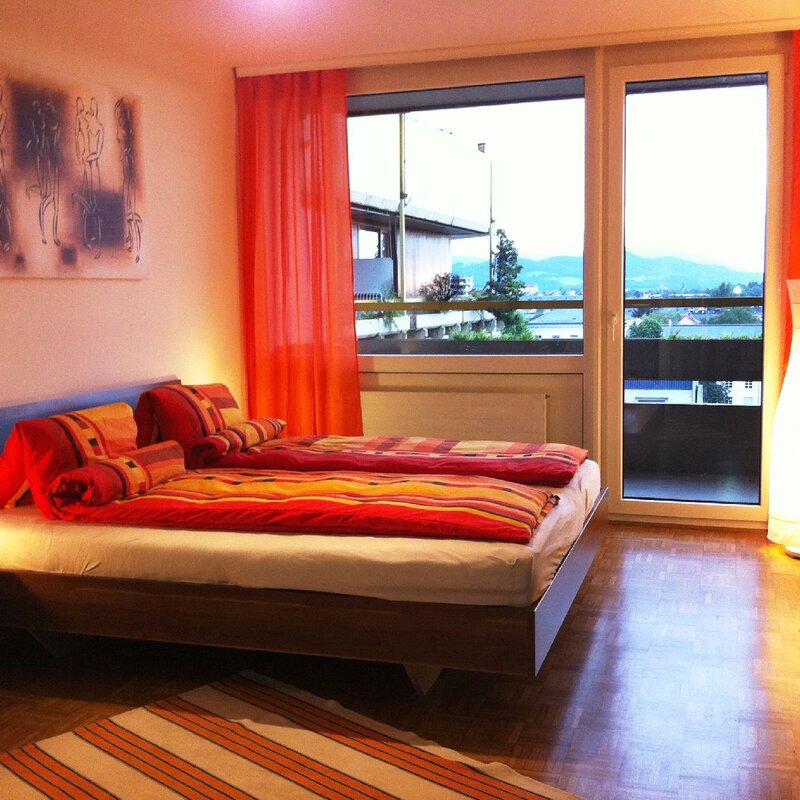 Dieses stilvolle Schlafzimmer einer möblierten Wohnung auf Zeit in der Steingasse, Salzburger Altstadt, überzeugt durch ein modernes Doppelbett mit hochwertigem Polster-Kopfteil und farbenfroher Bettwäsche in warmen Rot- und Orangetönen. Der gepflegte Parkettboden verleiht dem Raum eine wohnliche Atmosphäre, während großzügige Bodenfenster mit raumhohen orangefarbenen Vorhängen reichlich Tageslicht hereinlassen und einen freien Ausblick ins Freie bieten. Zwei stimmungsvolle Nachttischleuchten sowie eine markante Design-Stehleuchte sorgen für angenehmes Ambiente. Ein Flat-TV auf einem dunklen Sideboard sowie ein großer Einbauschrank runden die Ausstattung ab. Ein gerahmtes Kunstwerk an der Wand und ein gestreifter Teppich setzen dekorative Akzente. Diese Übergangswohnung eignet sich ideal für Berufspendler, Expats und Projektmitarbeiter, die in Salzburg für 1–6 Monate wohnen möchten. Zentrale Lage in der Salzburger Altstadt, vollmöbliert, flexibel buchbar auf kurzzeitmiete.at – ohne versteckte Kosten.