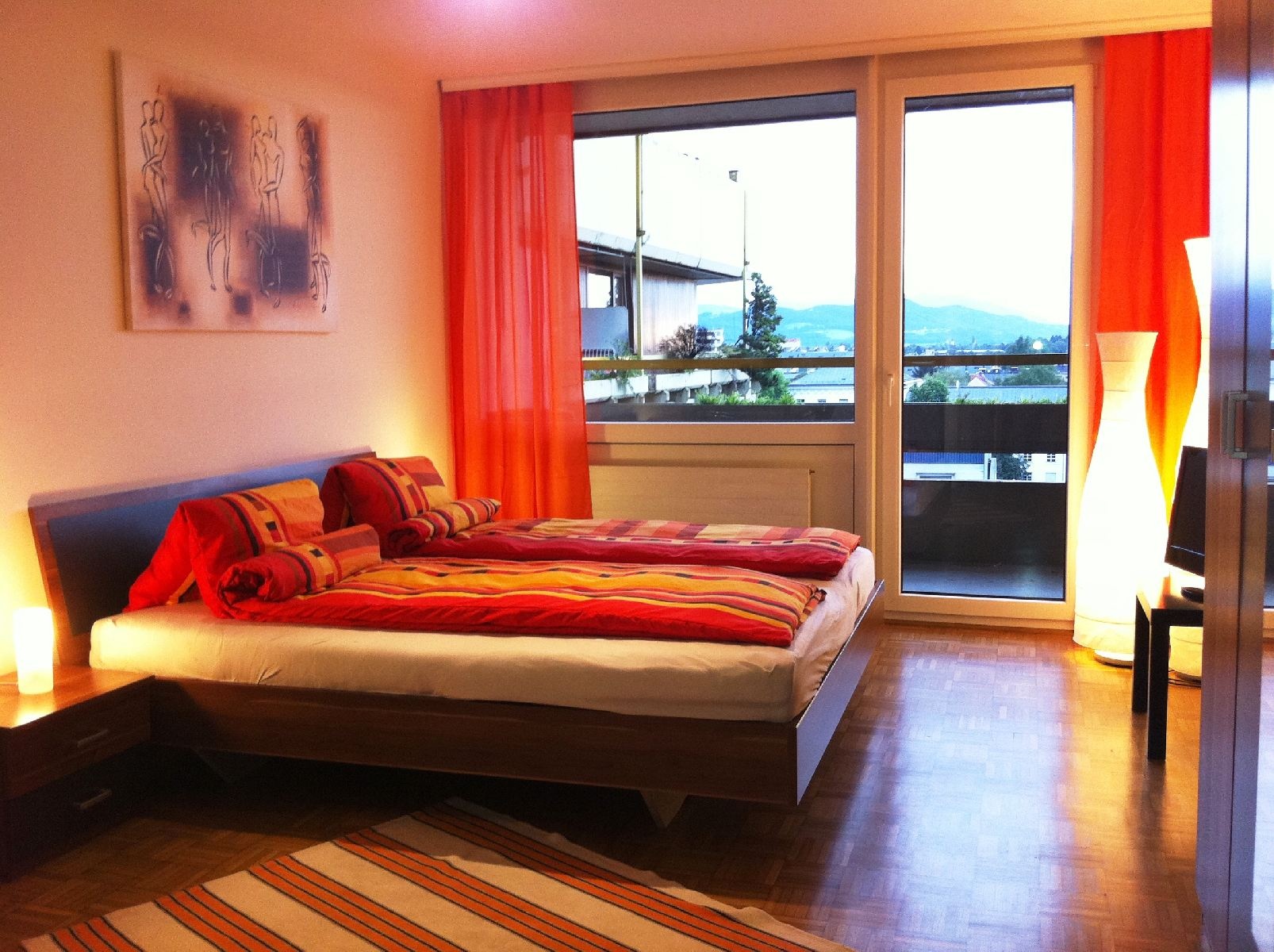 Dieses stilvolle Schlafzimmer einer möblierten Wohnung auf Zeit in der Steingasse, Salzburger Altstadt, überzeugt durch ein modernes Doppelbett mit hochwertigem Polster-Kopfteil und farbenfroher Bettwäsche in warmen Rot- und Orangetönen. Der gepflegte Parkettboden verleiht dem Raum eine wohnliche Atmosphäre, während großzügige Bodenfenster mit raumhohen orangefarbenen Vorhängen reichlich Tageslicht hereinlassen und einen freien Ausblick ins Freie bieten. Zwei stimmungsvolle Nachttischleuchten sowie eine markante Design-Stehleuchte sorgen für angenehmes Ambiente. Ein Flat-TV auf einem dunklen Sideboard sowie ein großer Einbauschrank runden die Ausstattung ab. Ein gerahmtes Kunstwerk an der Wand und ein gestreifter Teppich setzen dekorative Akzente. Diese Übergangswohnung eignet sich ideal für Berufspendler, Expats und Projektmitarbeiter, die in Salzburg für 1–6 Monate wohnen möchten. Zentrale Lage in der Salzburger Altstadt, vollmöbliert, flexibel buchbar auf kurzzeitmiete.at – ohne versteckte Kosten.