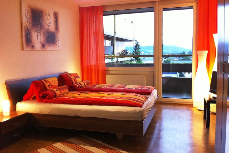 Dieses stilvolle Schlafzimmer einer möblierten Wohnung auf Zeit in der Steingasse, Salzburger Altstadt, überzeugt durch ein modernes Doppelbett mit hochwertigem Polster-Kopfteil und farbenfroher Bettwäsche in warmen Rot- und Orangetönen. Der gepflegte Parkettboden verleiht dem Raum eine wohnliche Atmosphäre, während großzügige Bodenfenster mit raumhohen orangefarbenen Vorhängen reichlich Tageslicht hereinlassen und einen freien Ausblick ins Freie bieten. Zwei stimmungsvolle Nachttischleuchten sowie eine markante Design-Stehleuchte sorgen für angenehmes Ambiente. Ein Flat-TV auf einem dunklen Sideboard sowie ein großer Einbauschrank runden die Ausstattung ab. Ein gerahmtes Kunstwerk an der Wand und ein gestreifter Teppich setzen dekorative Akzente. Diese Übergangswohnung eignet sich ideal für Berufspendler, Expats und Projektmitarbeiter, die in Salzburg für 1–6 Monate wohnen möchten. Zentrale Lage in der Salzburger Altstadt, vollmöbliert, flexibel buchbar auf kurzzeitmiete.at – ohne versteckte Kosten.
