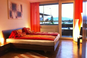 This stylish bedroom in a fully furnished short-term rental apartment on Steingasse in Salzburg Old Town features a modern double bed with an upholstered headboard and vibrant red and orange bedding. The well-maintained parquet flooring adds a warm, homely feel, while large floor-to-ceiling windows with bold orange curtains flood the room with natural light and offer open views to the outside. Two bedside lamps and a distinctive designer floor lamp create a welcoming ambiance. A flat-screen TV on a dark sideboard and a large built-in wardrobe complete the practical furnishings. A framed artwork on the wall and a striped area rug add tasteful decorative touches. Located in Salzburg's UNESCO World Heritage Old Town, this temporary living solution is perfectly suited for business travellers, expats, and consultants seeking furnished accommodation for 1–6 months. Centrally located, flexibly bookable via kurzzeitmiete.at – fully furnished, all-inclusive, no hidden costs.