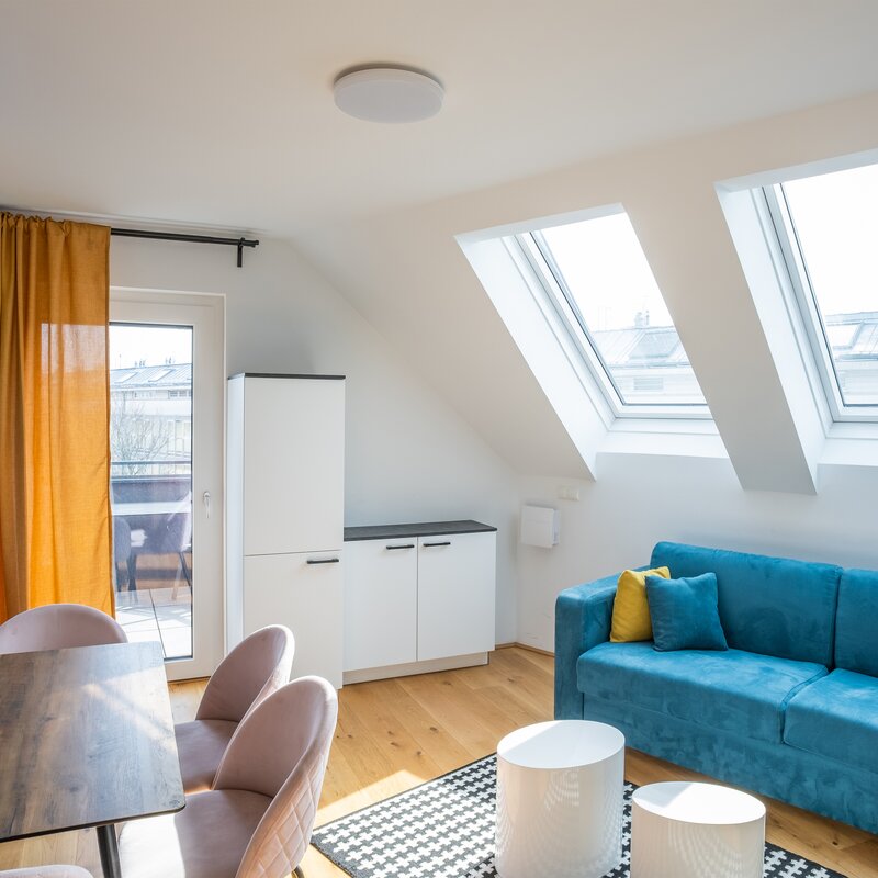 This furnished apartment for temporary living on Steingasse in Salzburg Old Town features a light-filled attic living room with two large skylights offering views across the rooftops of the UNESCO World Heritage city. The living area is equipped with a high-quality blue velvet sofa with yellow accent cushions, a dining table with soft rose upholstered chairs, and two round white coffee tables on a graphic-patterned rug. A wall-mounted flat-screen TV, a vibrant colourful art-print wall mural, and warm orange curtains create a modern, welcoming atmosphere. Light hardwood flooring, white built-in storage units, and direct access to an outdoor area complete the setup. This short-term rental is ideally suited for business travellers, expats, consultants, and project staff seeking a fully furnished temporary residence in central Salzburg for 1–6 months. Available via kurzzeitmiete.at – centrally located, flexibly bookable, all-inclusive, and with no hidden costs.