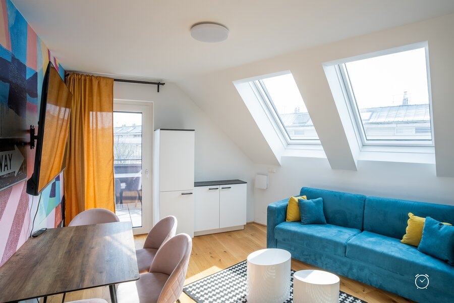 This furnished apartment for temporary living on Steingasse in Salzburg Old Town features a light-filled attic living room with two large skylights offering views across the rooftops of the UNESCO World Heritage city. The living area is equipped with a high-quality blue velvet sofa with yellow accent cushions, a dining table with soft rose upholstered chairs, and two round white coffee tables on a graphic-patterned rug. A wall-mounted flat-screen TV, a vibrant colourful art-print wall mural, and warm orange curtains create a modern, welcoming atmosphere. Light hardwood flooring, white built-in storage units, and direct access to an outdoor area complete the setup. This short-term rental is ideally suited for business travellers, expats, consultants, and project staff seeking a fully furnished temporary residence in central Salzburg for 1–6 months. Available via kurzzeitmiete.at – centrally located, flexibly bookable, all-inclusive, and with no hidden costs.