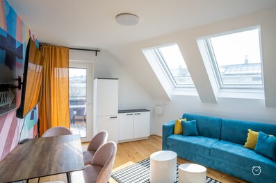 This furnished apartment for temporary living on Steingasse in Salzburg Old Town features a light-filled attic living room with two large skylights offering views across the rooftops of the UNESCO World Heritage city. The living area is equipped with a high-quality blue velvet sofa with yellow accent cushions, a dining table with soft rose upholstered chairs, and two round white coffee tables on a graphic-patterned rug. A wall-mounted flat-screen TV, a vibrant colourful art-print wall mural, and warm orange curtains create a modern, welcoming atmosphere. Light hardwood flooring, white built-in storage units, and direct access to an outdoor area complete the setup. This short-term rental is ideally suited for business travellers, expats, consultants, and project staff seeking a fully furnished temporary residence in central Salzburg for 1–6 months. Available via kurzzeitmiete.at – centrally located, flexibly bookable, all-inclusive, and with no hidden costs.
