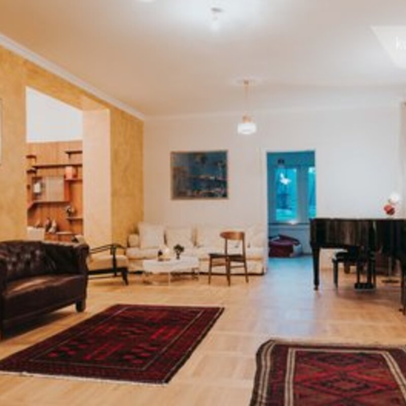 This furnished apartment for temporary living on Steingasse in Salzburg Old Town features a generously sized, light-filled living room with distinctive period charm. The bright parquet flooring, warm gold-toned walls, and impressive ceiling height create an elegant yet comfortable atmosphere. The room is visibly equipped with a classic dark leather Chesterfield sofa, additional seating, a grand piano, and several artworks adorning the walls. Two high-quality oriental rugs define the open living space, while a bookshelf and an adjoining room suggest a spacious overall floor plan. This short-term rental is ideally suited for business travellers, expats, and consultants seeking furnished accommodation in Salzburg for 1–6 months. Located in the UNESCO World Heritage zone of Salzburg Old Town, this temporary living solution on kurzzeitmiete.at offers full flexibility, transparent pricing, and no hidden costs – perfect for professionals who need a refined home base in the heart of Salzburg.