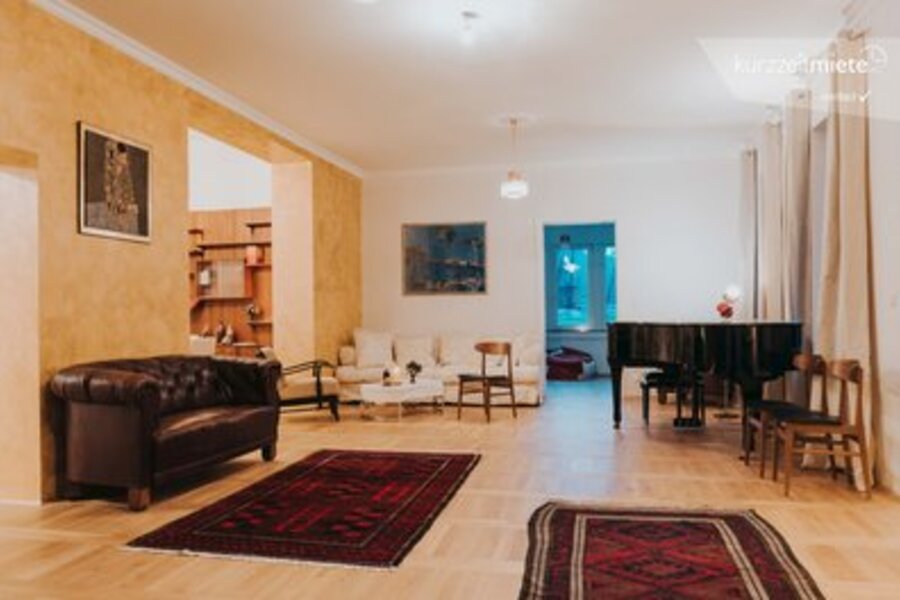 This furnished apartment for temporary living on Steingasse in Salzburg Old Town features a generously sized, light-filled living room with distinctive period charm. The bright parquet flooring, warm gold-toned walls, and impressive ceiling height create an elegant yet comfortable atmosphere. The room is visibly equipped with a classic dark leather Chesterfield sofa, additional seating, a grand piano, and several artworks adorning the walls. Two high-quality oriental rugs define the open living space, while a bookshelf and an adjoining room suggest a spacious overall floor plan. This short-term rental is ideally suited for business travellers, expats, and consultants seeking furnished accommodation in Salzburg for 1–6 months. Located in the UNESCO World Heritage zone of Salzburg Old Town, this temporary living solution on kurzzeitmiete.at offers full flexibility, transparent pricing, and no hidden costs – perfect for professionals who need a refined home base in the heart of Salzburg.