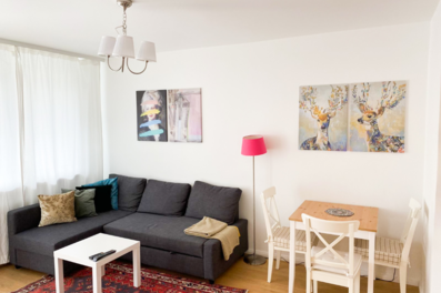 This bright and stylishly furnished living room is part of a temporary apartment available for short-term rental in Steingasse, Salzburg Old Town – a UNESCO World Heritage location. The space features a spacious dark grey corner sofa with decorative cushions and a cosy throw blanket, a white coffee table with a TV remote, and a compact wooden dining table with two white country-style chairs. A bold pink floor lamp adds a modern accent alongside carefully selected wall art, including a pop-art portrait and a decorative deer diptych. Natural light streams through full-length curtains, complemented by warm parquet flooring and a classic oriental rug. An elegant three-shade pendant light completes the welcoming atmosphere. Designed for business travellers, expats, consultants, and project workers, this furnished apartment is available for 1–6 months via kurzzeitmiete.at – centrally located, flexibly bookable, and transparently priced with no hidden costs.