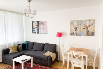 Dieses lichtdurchflutete Wohnzimmer einer möblierten Wohnung auf Zeit in der Steingasse, Salzburger Altstadt, überzeugt mit durchdachter Ausstattung für Berufspendler, Expats und Geschäftsreisende. Sichtbar sind ein großzügiges Ecksofa in Dunkelgrau mit dekorativen Kissen und einer Wolldecke, ein weißer Couchtisch mit TV-Fernbedienung sowie ein kleiner Esstisch aus Holz mit zwei weißen Stühlen im Landhausstil. Eine auffällige pinke Stehlampe setzt Akzente neben stimmungsvollen Kunstdrucken an der Wand – darunter ein Pop-Art-Motiv und ein dekoratives Hirsch-Diptychon. Natürliches Licht fällt durch bodenlange Vorhänge, der Boden ist mit Parkettdielen und einem klassischen Orientteppich gestaltet. Eine elegante Pendelleuchte mit drei Schirmen rundet das Ambiente ab. Diese Übergangswohnung steht für Kurzzeitmiete von 1–6 Monaten bereit – zentral gelegen im UNESCO-Weltkulturerbe Salzburg, flexibel buchbar und ohne versteckte Kosten auf kurzzeitmiete.at.