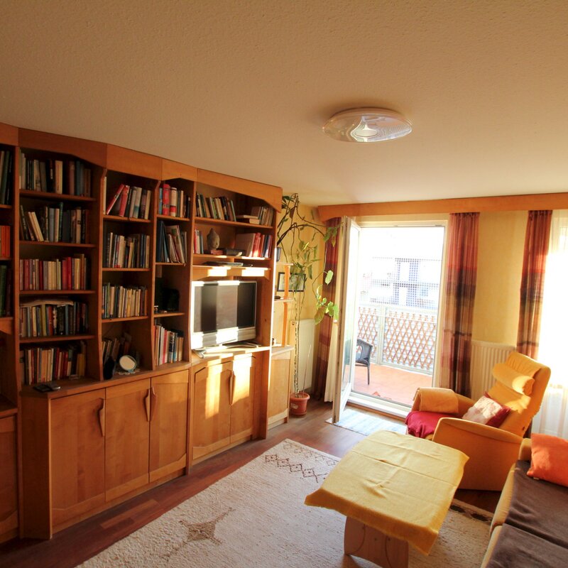 Bright, fully furnished living room available for short-term rental in the Steingasse, Salzburg Old Town – a UNESCO World Heritage location. The spacious room features a full-height solid wood bookshelf unit with an extensive book collection, a flat-screen TV integrated into the shelving system, and a stereo unit. Dark-stained hardwood flooring and warm wood tones throughout create a comfortable, home-like atmosphere ideal for temporary living. A large sliding glass door leads to a balcony terrace, flooding the room with natural light. The seating area includes a sofa and armchair with orange accent cushions, complemented by a central coffee table. A decorative indoor plant and ceiling light complete the furnishings. This furnished apartment is perfectly suited for business travellers, expats, and consultants seeking a fully equipped home in Salzburg Old Town for 1–6 months. Central location, flexible rental periods, and fully transparent pricing – no hidden costs, no compromises.
