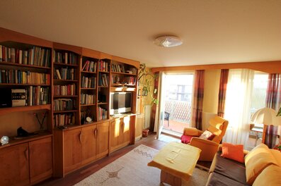 Bright, fully furnished living room available for short-term rental in the Steingasse, Salzburg Old Town – a UNESCO World Heritage location. The spacious room features a full-height solid wood bookshelf unit with an extensive book collection, a flat-screen TV integrated into the shelving system, and a stereo unit. Dark-stained hardwood flooring and warm wood tones throughout create a comfortable, home-like atmosphere ideal for temporary living. A large sliding glass door leads to a balcony terrace, flooding the room with natural light. The seating area includes a sofa and armchair with orange accent cushions, complemented by a central coffee table. A decorative indoor plant and ceiling light complete the furnishings. This furnished apartment is perfectly suited for business travellers, expats, and consultants seeking a fully equipped home in Salzburg Old Town for 1–6 months. Central location, flexible rental periods, and fully transparent pricing – no hidden costs, no compromises.