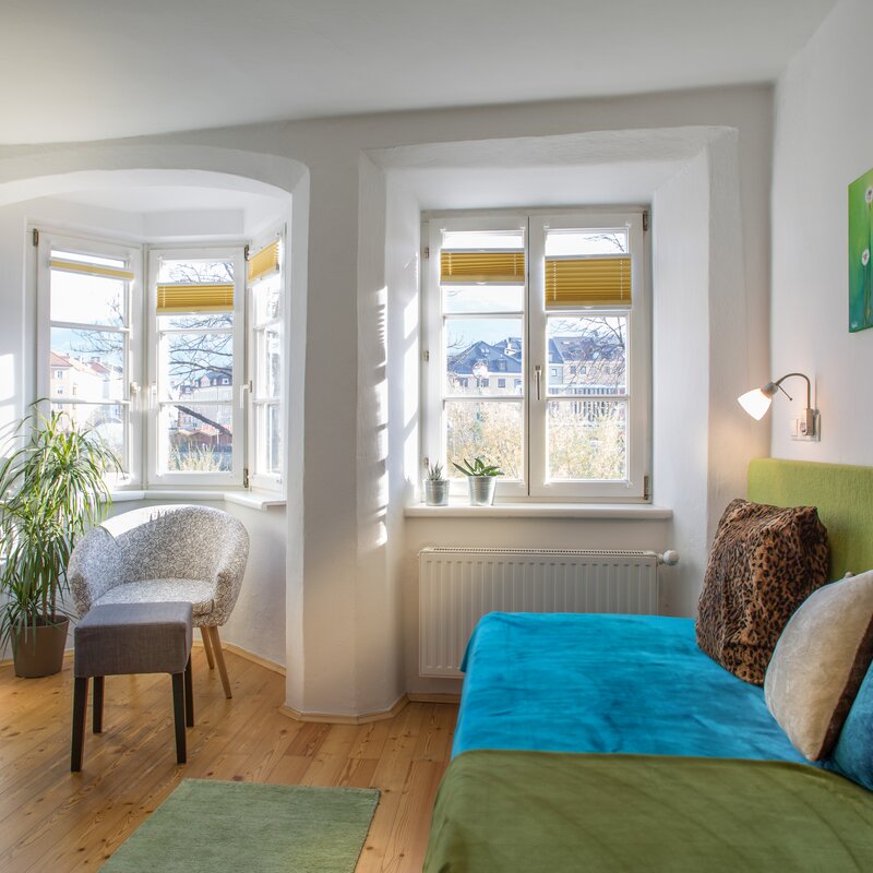 This sun-filled studio apartment in Salzburg Old Town – Steingasse – combines authentic Altbau character with modern comfort, making it an ideal short-term rental for business travellers, expats and project-based professionals. Clearly visible in the image: original hardwood plank flooring, white plastered walls with a charming arched alcove, large multi-pane casement windows with pleated blinds offering open city views. The furnished apartment features a comfortable sofa bed with teal upholstery and a green padded headboard, decorative cushions, and a wall-mounted reading lamp. Additional amenities include a flat-screen TV on a glass stand, a shelving unit with speakers, a lounge chair with matching footstool, a side table, and several indoor plants. A radiator beneath the window ensures year-round warmth. Two vibrant green art prints add a modern accent. Located in Salzburg's UNESCO World Heritage Old Town, this temporary living solution is available for 1–6 months. Move in ready, fully equipped, centrally located – no hidden costs, no hassle.