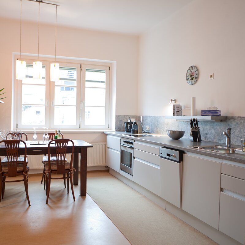This bright and well-appointed eat-in kitchen is ideal for business travellers, expats, and consultants seeking furnished temporary living in Salzburg Old Town, Steingasse. The fully equipped fitted kitchen features a stainless steel worktop, ceramic hob, oven, dishwasher, and a full-size fridge-freezer – everything needed for comfortable short-term rental living. A natural stone tile splashback and under-cabinet lighting add a premium feel, while a knife block set and coffee machine ensure practical day-to-day convenience. The solid wood dining table with four classic Thonet-style chairs seats up to four guests, lit by elegant pendant lamps that create a welcoming atmosphere. An antique sideboard with mirror reflects the historic character of this old-building apartment. Large double windows flood the space with natural daylight. Located in the UNESCO World Heritage Site of Salzburg Old Town, this furnished apartment is available for 1–6 months – centrally situated, flexibly rentable, and transparent in pricing with no hidden costs. Perfect for professionals relocating or working on short-term assignments in Salzburg.