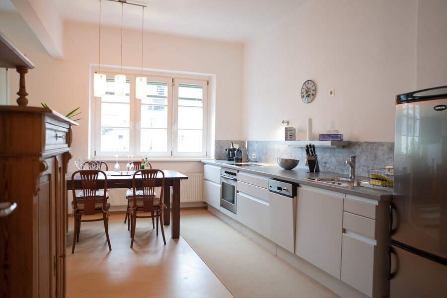 This bright and well-appointed eat-in kitchen is ideal for business travellers, expats, and consultants seeking furnished temporary living in Salzburg Old Town, Steingasse. The fully equipped fitted kitchen features a stainless steel worktop, ceramic hob, oven, dishwasher, and a full-size fridge-freezer – everything needed for comfortable short-term rental living. A natural stone tile splashback and under-cabinet lighting add a premium feel, while a knife block set and coffee machine ensure practical day-to-day convenience. The solid wood dining table with four classic Thonet-style chairs seats up to four guests, lit by elegant pendant lamps that create a welcoming atmosphere. An antique sideboard with mirror reflects the historic character of this old-building apartment. Large double windows flood the space with natural daylight. Located in the UNESCO World Heritage Site of Salzburg Old Town, this furnished apartment is available for 1–6 months – centrally situated, flexibly rentable, and transparent in pricing with no hidden costs. Perfect for professionals relocating or working on short-term assignments in Salzburg.