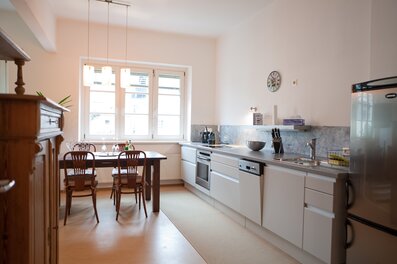 This bright and well-appointed eat-in kitchen is ideal for business travellers, expats, and consultants seeking furnished temporary living in Salzburg Old Town, Steingasse. The fully equipped fitted kitchen features a stainless steel worktop, ceramic hob, oven, dishwasher, and a full-size fridge-freezer – everything needed for comfortable short-term rental living. A natural stone tile splashback and under-cabinet lighting add a premium feel, while a knife block set and coffee machine ensure practical day-to-day convenience. The solid wood dining table with four classic Thonet-style chairs seats up to four guests, lit by elegant pendant lamps that create a welcoming atmosphere. An antique sideboard with mirror reflects the historic character of this old-building apartment. Large double windows flood the space with natural daylight. Located in the UNESCO World Heritage Site of Salzburg Old Town, this furnished apartment is available for 1–6 months – centrally situated, flexibly rentable, and transparent in pricing with no hidden costs. Perfect for professionals relocating or working on short-term assignments in Salzburg.