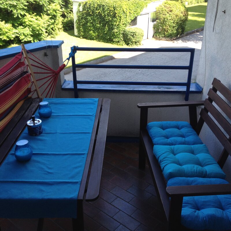 This furnished apartment for temporary living on Steingasse in Salzburg Old Town – a UNESCO World Heritage site – features a spacious covered balcony with a complete solid wood outdoor furniture set: a rectangular dining table with a blue textile table runner, a sturdy wooden bench, and a two-seater garden sofa with comfortable turquoise cushions. Decorative candle holders and an ashtray in blue tones are neatly arranged on the table. A vibrant red-and-yellow hammock chair adds a relaxed touch to the outdoor space. The blue-painted metal railing opens onto a well-maintained green area with lawn, hedges, and mature trees – perfect for unwinding after a long workday. The terracotta-style tiled floor lends a warm, Mediterranean character to the balcony. This short-term rental is ideally suited for business travellers, expats, and consultants seeking flexible furnished accommodation in Salzburg Old Town for 1–6 months. Central location, transparent pricing – no hidden costs.