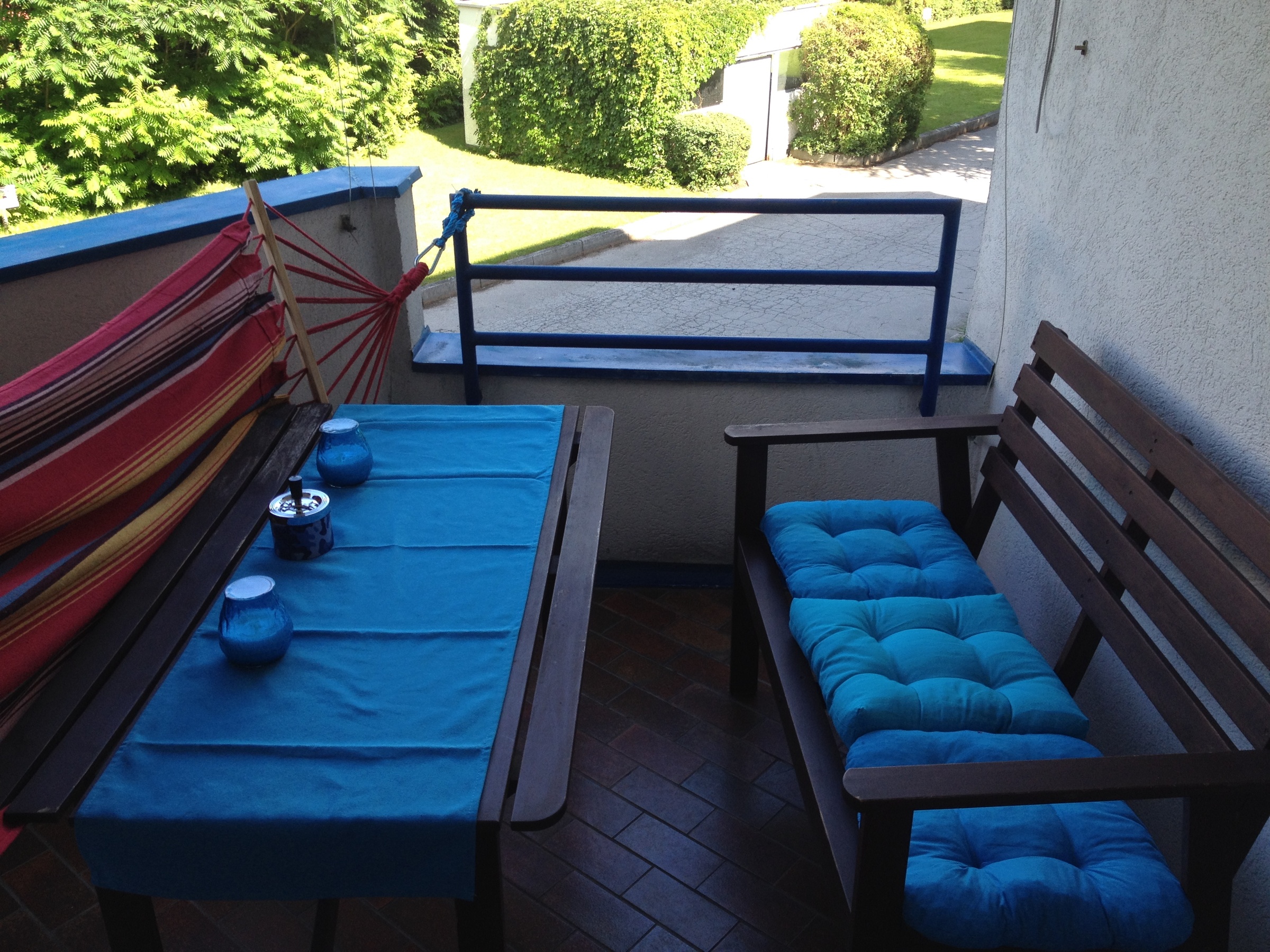 This furnished apartment for temporary living on Steingasse in Salzburg Old Town – a UNESCO World Heritage site – features a spacious covered balcony with a complete solid wood outdoor furniture set: a rectangular dining table with a blue textile table runner, a sturdy wooden bench, and a two-seater garden sofa with comfortable turquoise cushions. Decorative candle holders and an ashtray in blue tones are neatly arranged on the table. A vibrant red-and-yellow hammock chair adds a relaxed touch to the outdoor space. The blue-painted metal railing opens onto a well-maintained green area with lawn, hedges, and mature trees – perfect for unwinding after a long workday. The terracotta-style tiled floor lends a warm, Mediterranean character to the balcony. This short-term rental is ideally suited for business travellers, expats, and consultants seeking flexible furnished accommodation in Salzburg Old Town for 1–6 months. Central location, transparent pricing – no hidden costs.