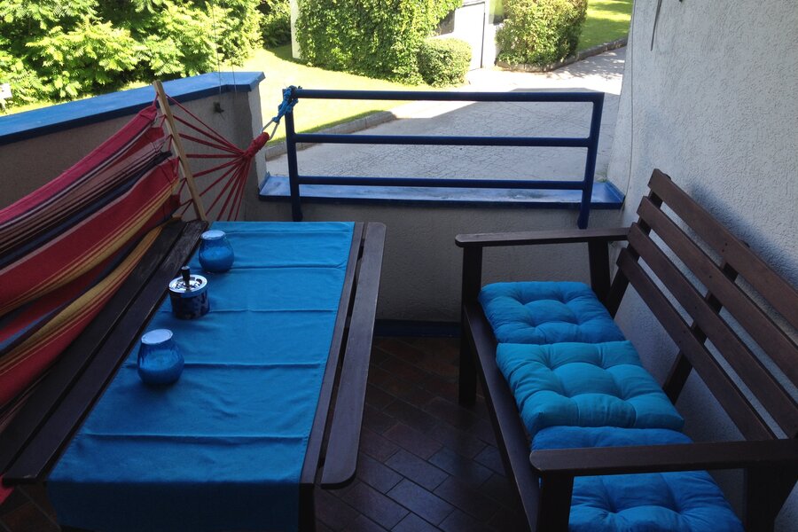 This furnished apartment for temporary living on Steingasse in Salzburg Old Town – a UNESCO World Heritage site – features a spacious covered balcony with a complete solid wood outdoor furniture set: a rectangular dining table with a blue textile table runner, a sturdy wooden bench, and a two-seater garden sofa with comfortable turquoise cushions. Decorative candle holders and an ashtray in blue tones are neatly arranged on the table. A vibrant red-and-yellow hammock chair adds a relaxed touch to the outdoor space. The blue-painted metal railing opens onto a well-maintained green area with lawn, hedges, and mature trees – perfect for unwinding after a long workday. The terracotta-style tiled floor lends a warm, Mediterranean character to the balcony. This short-term rental is ideally suited for business travellers, expats, and consultants seeking flexible furnished accommodation in Salzburg Old Town for 1–6 months. Central location, transparent pricing – no hidden costs.