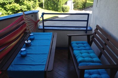This furnished apartment for temporary living on Steingasse in Salzburg Old Town – a UNESCO World Heritage site – features a spacious covered balcony with a complete solid wood outdoor furniture set: a rectangular dining table with a blue textile table runner, a sturdy wooden bench, and a two-seater garden sofa with comfortable turquoise cushions. Decorative candle holders and an ashtray in blue tones are neatly arranged on the table. A vibrant red-and-yellow hammock chair adds a relaxed touch to the outdoor space. The blue-painted metal railing opens onto a well-maintained green area with lawn, hedges, and mature trees – perfect for unwinding after a long workday. The terracotta-style tiled floor lends a warm, Mediterranean character to the balcony. This short-term rental is ideally suited for business travellers, expats, and consultants seeking flexible furnished accommodation in Salzburg Old Town for 1–6 months. Central location, transparent pricing – no hidden costs.