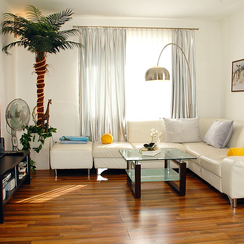 This sun-filled living room of a furnished apartment for temporary living on Steingasse in Salzburg's UNESCO World Heritage Old Town offers everything business travellers, expats, and consultants need for a comfortable stay. Clearly visible are a spacious white leather corner sofa with decorative cushions, a contemporary glass coffee table with a dark wood frame, and a flat-screen TV on a sleek black media unit with storage shelves. The warm-toned hardwood parquet floor adds an upscale feel throughout the space. A curved silver arc floor lamp provides elegant ambient lighting, complemented by floor-length silver curtains framing the bright windows. Lush greenery – including a decorative palm tree with fairy lights and a spider plant – alongside a desk fan and a white orchid create a welcoming, homely atmosphere. A framed artwork on the wall completes the modern interior. Available for short-term rental from 1 to 6 months on kurzzeitmiete.at – centrally located, fully furnished, and transparently priced with no hidden costs.