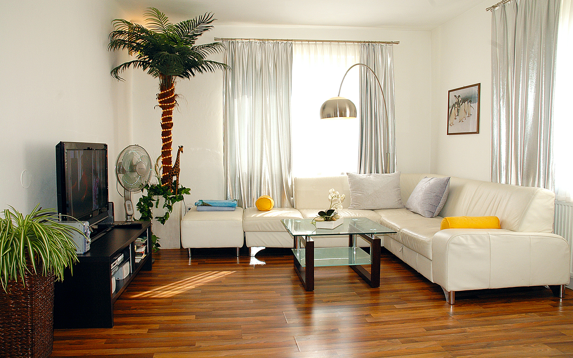 This sun-filled living room of a furnished apartment for temporary living on Steingasse in Salzburg's UNESCO World Heritage Old Town offers everything business travellers, expats, and consultants need for a comfortable stay. Clearly visible are a spacious white leather corner sofa with decorative cushions, a contemporary glass coffee table with a dark wood frame, and a flat-screen TV on a sleek black media unit with storage shelves. The warm-toned hardwood parquet floor adds an upscale feel throughout the space. A curved silver arc floor lamp provides elegant ambient lighting, complemented by floor-length silver curtains framing the bright windows. Lush greenery – including a decorative palm tree with fairy lights and a spider plant – alongside a desk fan and a white orchid create a welcoming, homely atmosphere. A framed artwork on the wall completes the modern interior. Available for short-term rental from 1 to 6 months on kurzzeitmiete.at – centrally located, fully furnished, and transparently priced with no hidden costs.