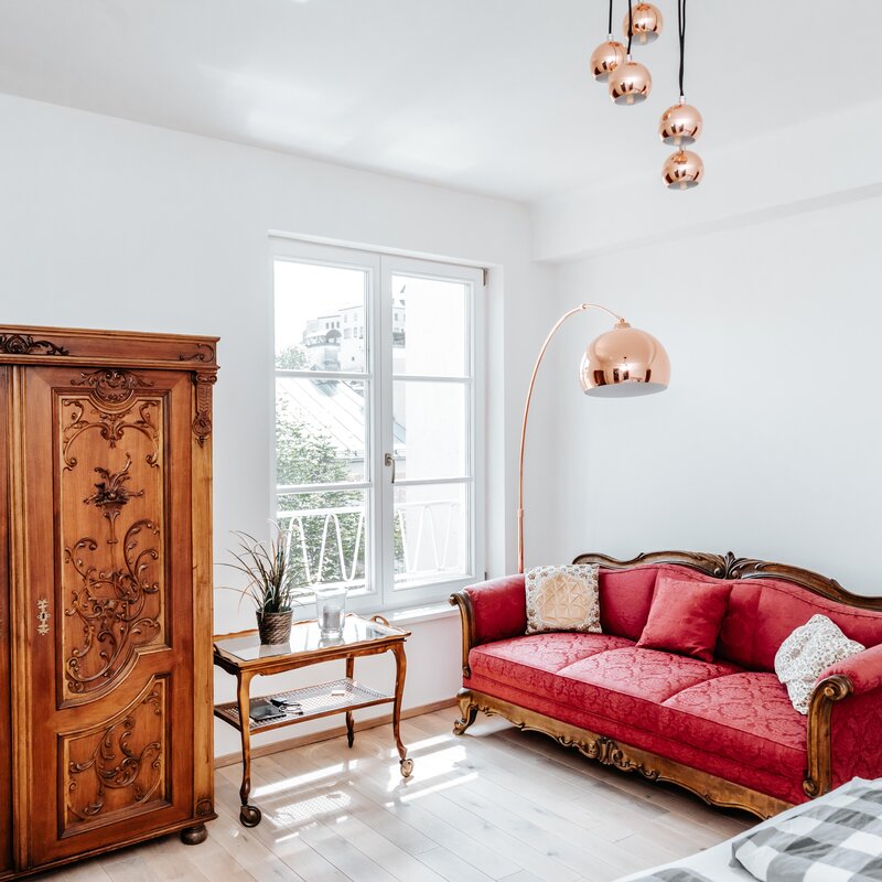 This furnished apartment for temporary living on Steingasse in Salzburg Old Town blends historic charm with contemporary design accents. The living room features a magnificent hand-carved baroque wardrobe and an elegant red damask sofa with ornately crafted wooden frames – both high-quality antique pieces. A delicate glass-top side table with curved legs complements the classic interior. Copper-toned pendant lights and a sculptural arc floor lamp in a rose-gold finish introduce modern style elements. The light parquet flooring and bright white walls create an airy, sun-filled atmosphere. A French-style balcony door with latticed windows opens onto an outdoor area with city views. A potted plant and glass tray table complete the refined setting. Perfect for business travellers, expats, and consultants seeking a short-term rental in Salzburg's UNESCO World Heritage zone for 1–6 months. Fully furnished, centrally located, and transparently priced – available via kurzzeitmiete.at.