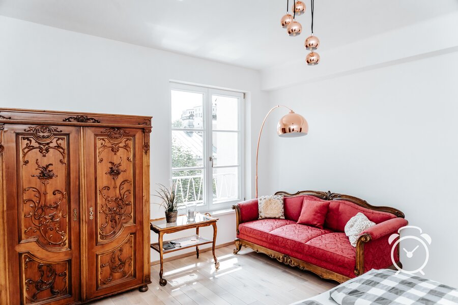 This furnished apartment for temporary living on Steingasse in Salzburg Old Town blends historic charm with contemporary design accents. The living room features a magnificent hand-carved baroque wardrobe and an elegant red damask sofa with ornately crafted wooden frames – both high-quality antique pieces. A delicate glass-top side table with curved legs complements the classic interior. Copper-toned pendant lights and a sculptural arc floor lamp in a rose-gold finish introduce modern style elements. The light parquet flooring and bright white walls create an airy, sun-filled atmosphere. A French-style balcony door with latticed windows opens onto an outdoor area with city views. A potted plant and glass tray table complete the refined setting. Perfect for business travellers, expats, and consultants seeking a short-term rental in Salzburg's UNESCO World Heritage zone for 1–6 months. Fully furnished, centrally located, and transparently priced – available via kurzzeitmiete.at.
