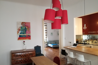Diese möblierte Wohnung auf Zeit in der Steingasse, Salzburger Altstadt, überzeugt mit einem großzügigen, offen gestalteten Wohn- und Küchenbereich im charaktervollen Altbau. Sichtbar sind ein massiver Holzesstisch mit Stühlen, eine vollausgestattete offene Küche mit roter Hochglanzfront, Gaskochfeld, Spüle und Küchenbar mit Barhockern. Ein auffälliges rotes Hängelampensystem sorgt für stimmungsvolles Ambiente. Die Ausstattung umfasst Parkettboden im Fischgrätmuster, eine antike Holzkommode, dekorative Kunstwerke an der Wand sowie einen sichtbaren Laptop-Arbeitsplatz an der Kücheninsel – ideal für mobiles Arbeiten. Durch den Durchgang ist ein weiterer Wohnbereich mit Sofa erkennbar. Die Deckenhöhe unterstreicht den typischen Altbau-Charme. Diese Übergangswohnung eignet sich perfekt für Berufspendler, Expats und Projektmitarbeiter, die eine vollmöblierte Unterkunft für 1–6 Monate in zentraler Lage der Salzburger Altstadt suchen. Kurzzeitmiete auf kurzzeitmiete.at – flexibel, zentral und transparent.