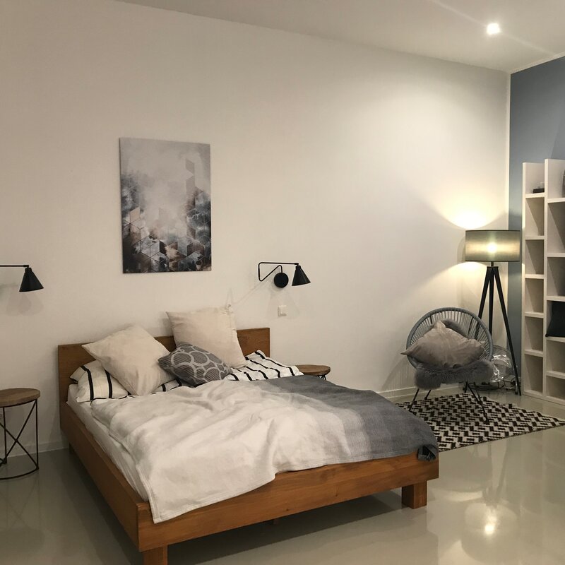 This furnished apartment for temporary living on Steingasse in Salzburg Old Town features a stylishly designed bedroom with a distinct Scandinavian aesthetic. The spacious double bed with a solid wooden frame is dressed with high-quality bedding and multiple pillows for maximum comfort. Two adjustable industrial-style wall-mounted reading lamps provide targeted lighting, complemented by a modern tripod floor lamp that adds warm ambient light. A round wooden side table and a wire-frame metal stool serve as practical surfaces on either side of the bed. An open white wall shelving unit offers storage and display space, while a two-tone steel-blue accent wall creates a contemporary visual contrast. The Acapulco-style lounge chair with a soft cushion and a black-and-white geometric area rug define a cosy seating corner. Located in UNESCO World Heritage-listed Salzburg Old Town, this short-term rental on Steingasse is ideal for business travellers, expats, and consultants seeking flexible temporary housing for 1–6 months – fully furnished, centrally located, and with no hidden costs.