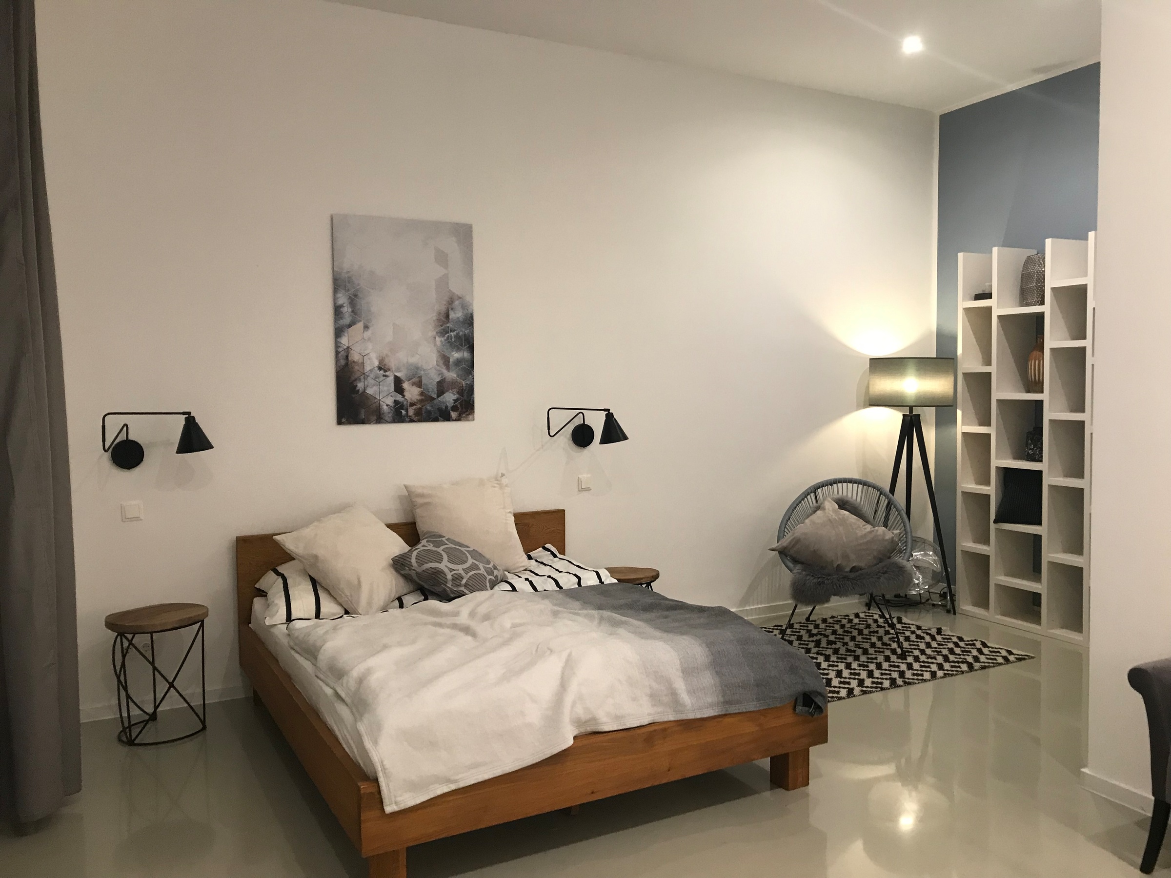 This furnished apartment for temporary living on Steingasse in Salzburg Old Town features a stylishly designed bedroom with a distinct Scandinavian aesthetic. The spacious double bed with a solid wooden frame is dressed with high-quality bedding and multiple pillows for maximum comfort. Two adjustable industrial-style wall-mounted reading lamps provide targeted lighting, complemented by a modern tripod floor lamp that adds warm ambient light. A round wooden side table and a wire-frame metal stool serve as practical surfaces on either side of the bed. An open white wall shelving unit offers storage and display space, while a two-tone steel-blue accent wall creates a contemporary visual contrast. The Acapulco-style lounge chair with a soft cushion and a black-and-white geometric area rug define a cosy seating corner. Located in UNESCO World Heritage-listed Salzburg Old Town, this short-term rental on Steingasse is ideal for business travellers, expats, and consultants seeking flexible temporary housing for 1–6 months – fully furnished, centrally located, and with no hidden costs.