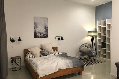 This furnished apartment for temporary living on Steingasse in Salzburg Old Town features a stylishly designed bedroom with a distinct Scandinavian aesthetic. The spacious double bed with a solid wooden frame is dressed with high-quality bedding and multiple pillows for maximum comfort. Two adjustable industrial-style wall-mounted reading lamps provide targeted lighting, complemented by a modern tripod floor lamp that adds warm ambient light. A round wooden side table and a wire-frame metal stool serve as practical surfaces on either side of the bed. An open white wall shelving unit offers storage and display space, while a two-tone steel-blue accent wall creates a contemporary visual contrast. The Acapulco-style lounge chair with a soft cushion and a black-and-white geometric area rug define a cosy seating corner. Located in UNESCO World Heritage-listed Salzburg Old Town, this short-term rental on Steingasse is ideal for business travellers, expats, and consultants seeking flexible temporary housing for 1–6 months – fully furnished, centrally located, and with no hidden costs.