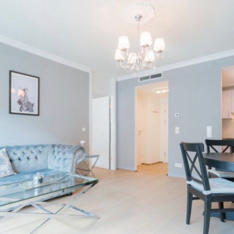 This furnished apartment for temporary living on Steingasse in Salzburg Old Town – a UNESCO World Heritage Site – features a tastefully designed open-plan living and dining area in calming light blue and grey tones. The living room is appointed with a tufted Chesterfield sofa in powder blue with decorative cushions, a contemporary glass coffee table on a chrome frame, and an elegant multi-arm chandelier. High ceilings with white crown moulding add a sense of refined, classic character to this short-term rental. The dining area offers a round dark-wood table with matching chairs for up to four guests. Through the connecting archway, a fully equipped white fitted kitchen is clearly visible. Black-and-white animal print wall art and light-toned flooring complete the bright, welcoming atmosphere. This furnished apartment is ideally suited for business travellers, expats, and consultants seeking quality temporary living in central Salzburg for 1–6 months – flexible lease terms, fully inclusive, and no hidden costs.