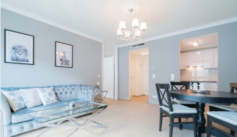 This furnished apartment for temporary living on Steingasse in Salzburg Old Town – a UNESCO World Heritage Site – features a tastefully designed open-plan living and dining area in calming light blue and grey tones. The living room is appointed with a tufted Chesterfield sofa in powder blue with decorative cushions, a contemporary glass coffee table on a chrome frame, and an elegant multi-arm chandelier. High ceilings with white crown moulding add a sense of refined, classic character to this short-term rental. The dining area offers a round dark-wood table with matching chairs for up to four guests. Through the connecting archway, a fully equipped white fitted kitchen is clearly visible. Black-and-white animal print wall art and light-toned flooring complete the bright, welcoming atmosphere. This furnished apartment is ideally suited for business travellers, expats, and consultants seeking quality temporary living in central Salzburg for 1–6 months – flexible lease terms, fully inclusive, and no hidden costs.