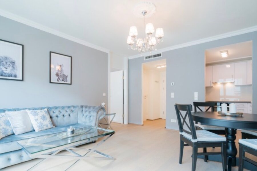This furnished apartment for temporary living on Steingasse in Salzburg Old Town – a UNESCO World Heritage Site – features a tastefully designed open-plan living and dining area in calming light blue and grey tones. The living room is appointed with a tufted Chesterfield sofa in powder blue with decorative cushions, a contemporary glass coffee table on a chrome frame, and an elegant multi-arm chandelier. High ceilings with white crown moulding add a sense of refined, classic character to this short-term rental. The dining area offers a round dark-wood table with matching chairs for up to four guests. Through the connecting archway, a fully equipped white fitted kitchen is clearly visible. Black-and-white animal print wall art and light-toned flooring complete the bright, welcoming atmosphere. This furnished apartment is ideally suited for business travellers, expats, and consultants seeking quality temporary living in central Salzburg for 1–6 months – flexible lease terms, fully inclusive, and no hidden costs.