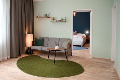 This furnished apartment for temporary living on Steingasse in Salzburg's UNESCO World Heritage Old Town features a thoughtfully designed living and sleeping area with a modern retro aesthetic. The living room showcases a quality three-seat sofa with wooden armrests and accent cushions, an organically shaped green shag rug, a mid-century walnut coffee table with gold-tipped legs, and warm ambient floor lighting. Oak wall shelves styled with books, plants, and decorative objects add a homely feel. Light parquet flooring and sage-green walls create a calm, contemporary atmosphere. Through the open doorway, a separate bedroom with a double bed, bedside lighting, and a deep navy accent wall is clearly visible – ideal for extended stays. Full-length blackout curtains in taupe ensure maximum privacy. This short-term rental in Salzburg Old Town is perfectly suited for business travellers, expats, and project consultants seeking flexible, fully furnished accommodation for 1–6 months – centrally located, all-inclusive, and with no hidden costs.