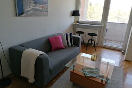 Wohnraum, ausgestattet mit einer Couch, einem Tisch, Theke am Fenster mit 2 Hockern, Schubladkasten, großer Kleiderschrank und einer Schlafnische mit einem Bett 140x200